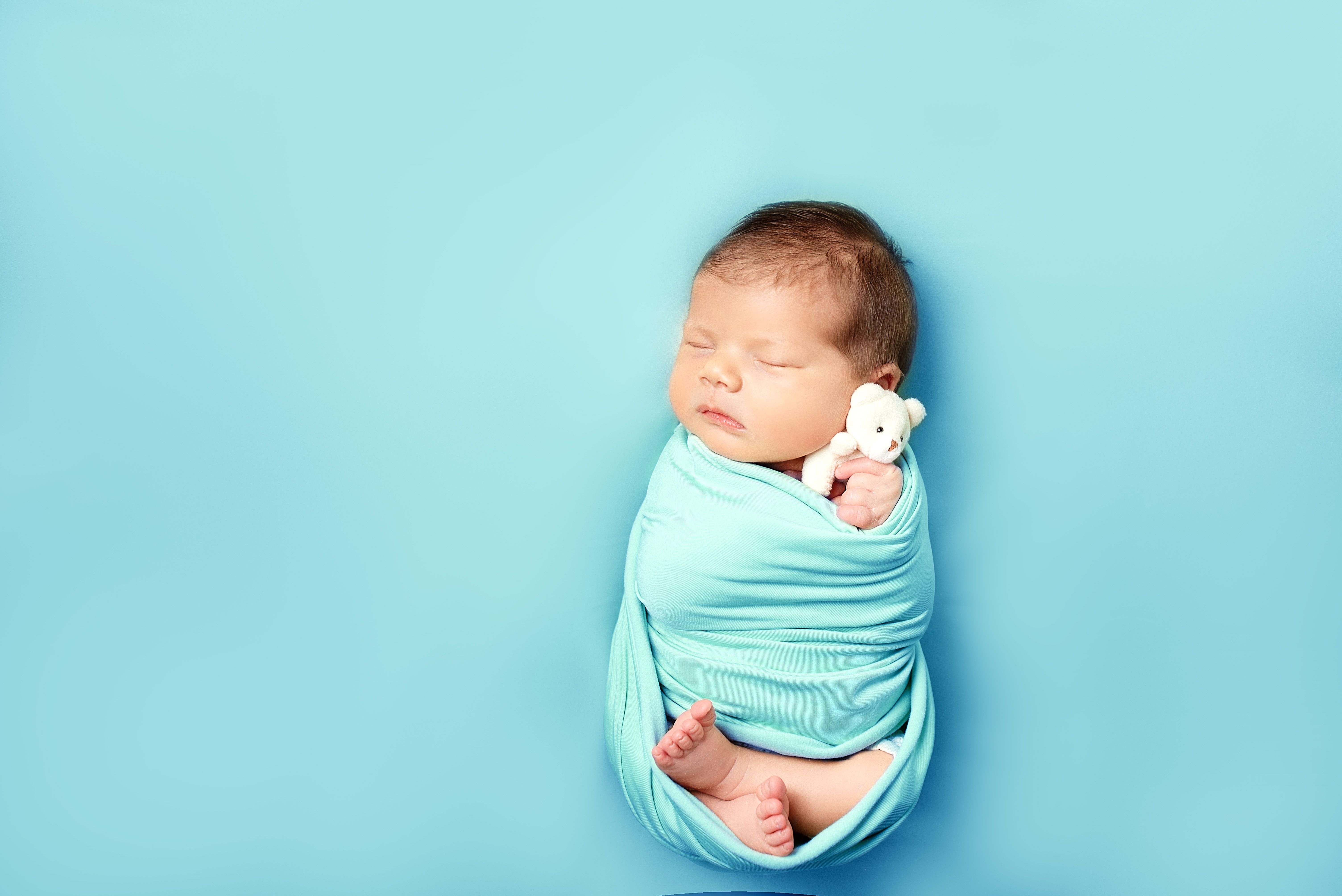 Adorable newborn baby wrapped in a blue blanket, holding a tiny teddy bear on a soft blue background.