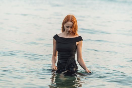 A woman with red hair wearing a black dress standing in a calm sea during the day.