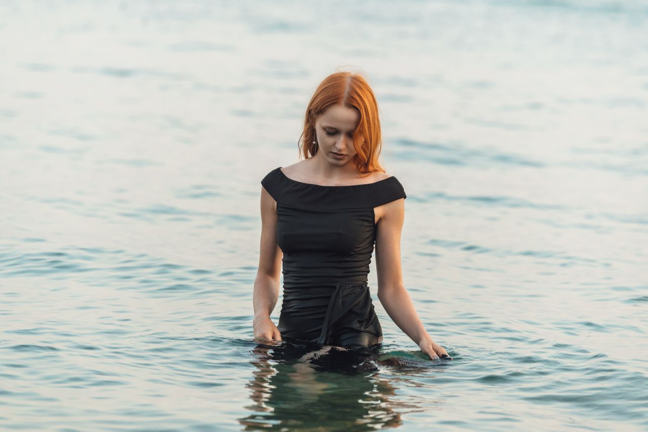 a woman with red hair wearing a black dress standing in a calm sea dur