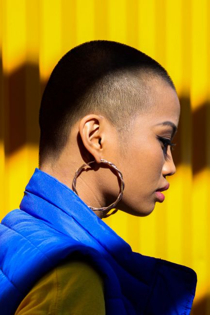 Bold profile portrait of a woman with shaved head and hoop earring against a vibrant yellow background.