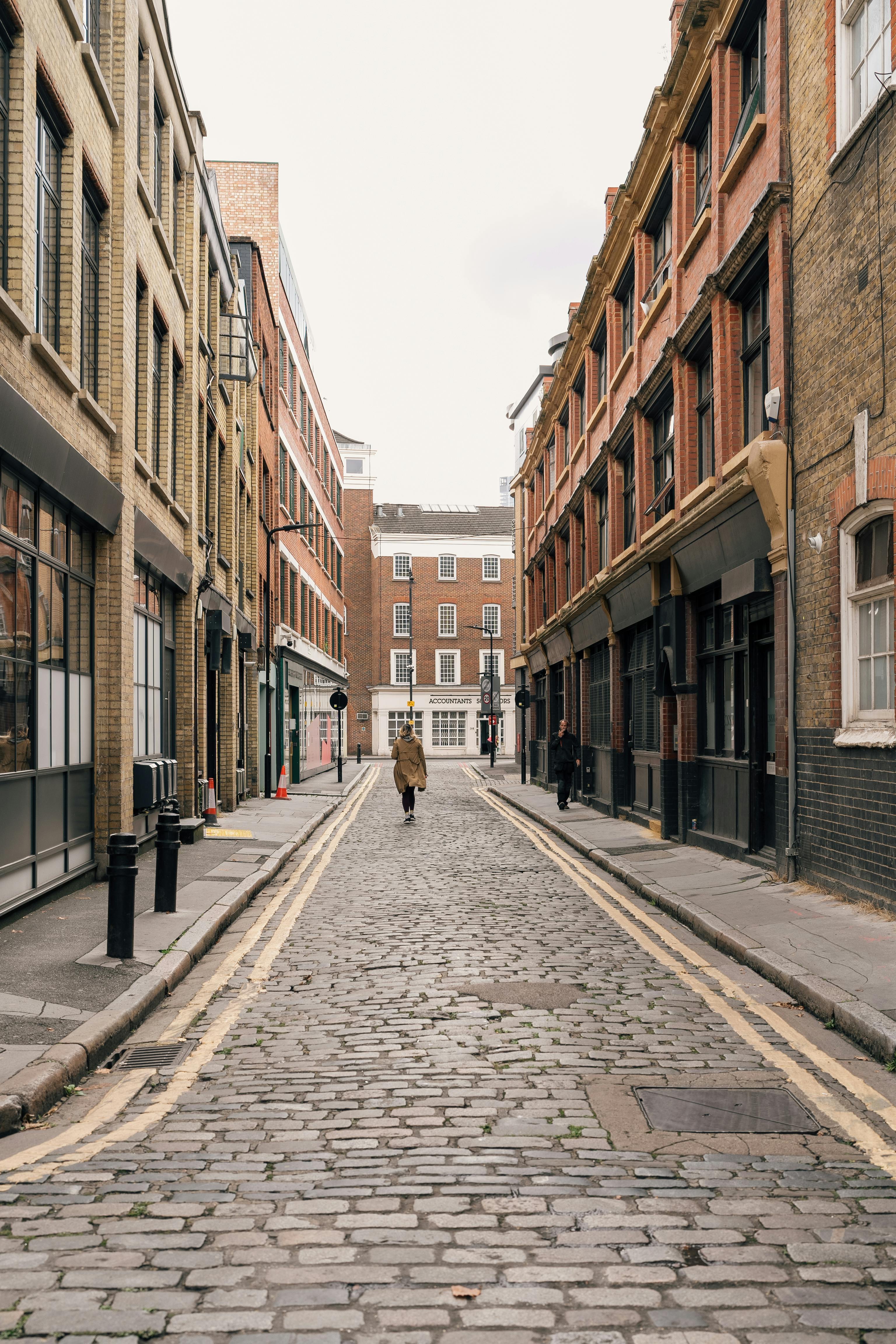 A solitary figure walks down a cobbled street lined with historic brick buildings in Londra, UK.