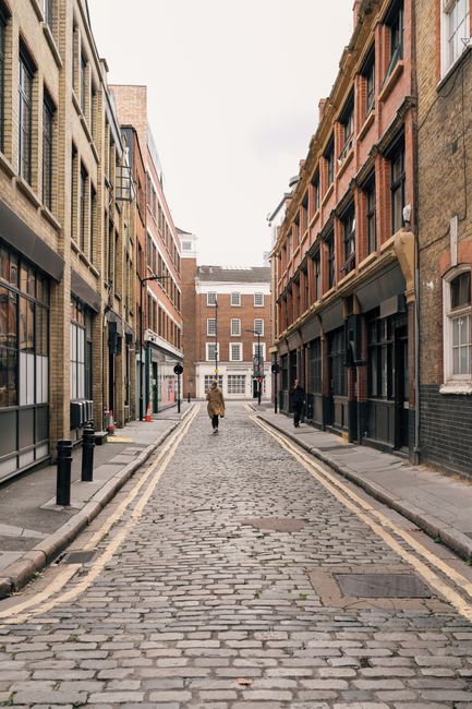 A solitary figure walks down a cobbled street lined with historic brick buildings in Londra, UK.