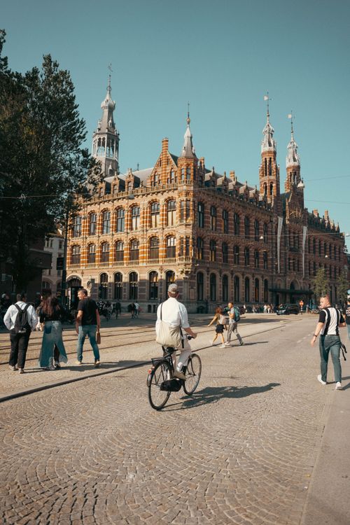 Free A cyclist rides past a historic building in a bustling European city, with pedestrians enjoying the sunny day. Stock Photo