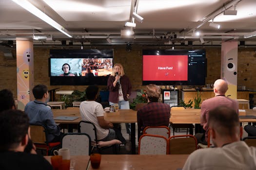A speaker engages with an audience in a modern conference setup. Interactive screens enhance the presentation.