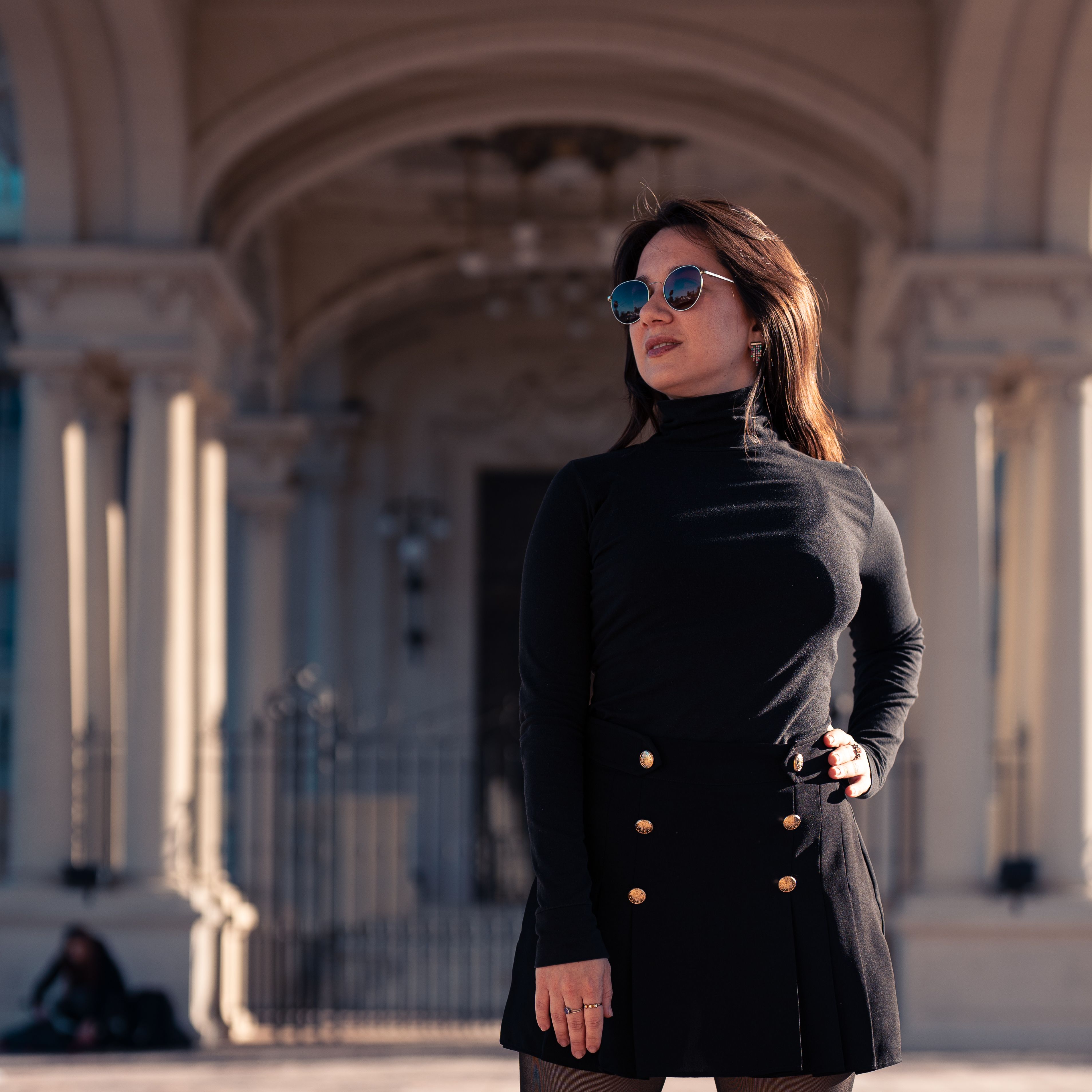 Confident woman in black outfit and sunglasses stands before grand architectural archway.