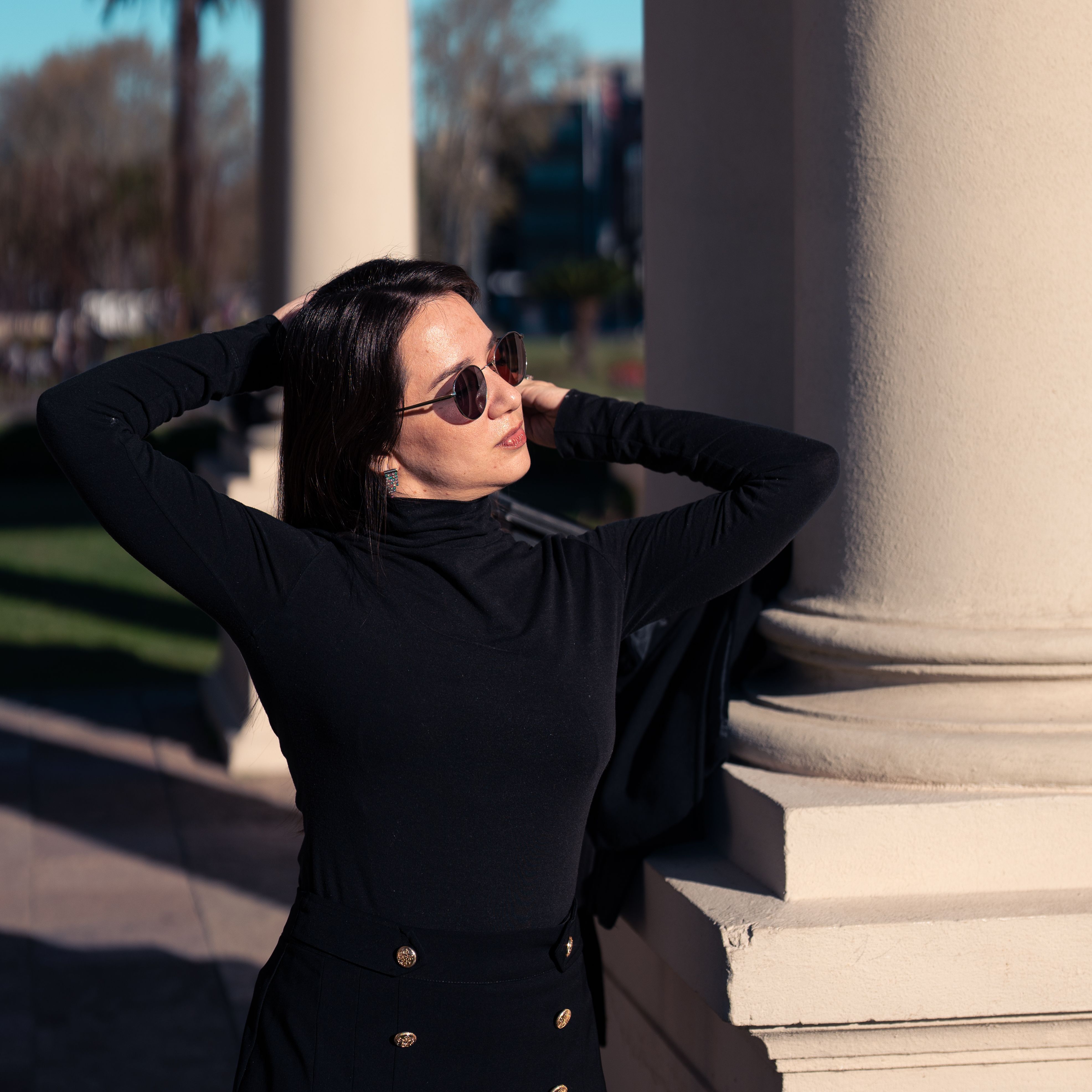 Stylish woman in dark outfit poses confidently in sunlight near grand pillars.