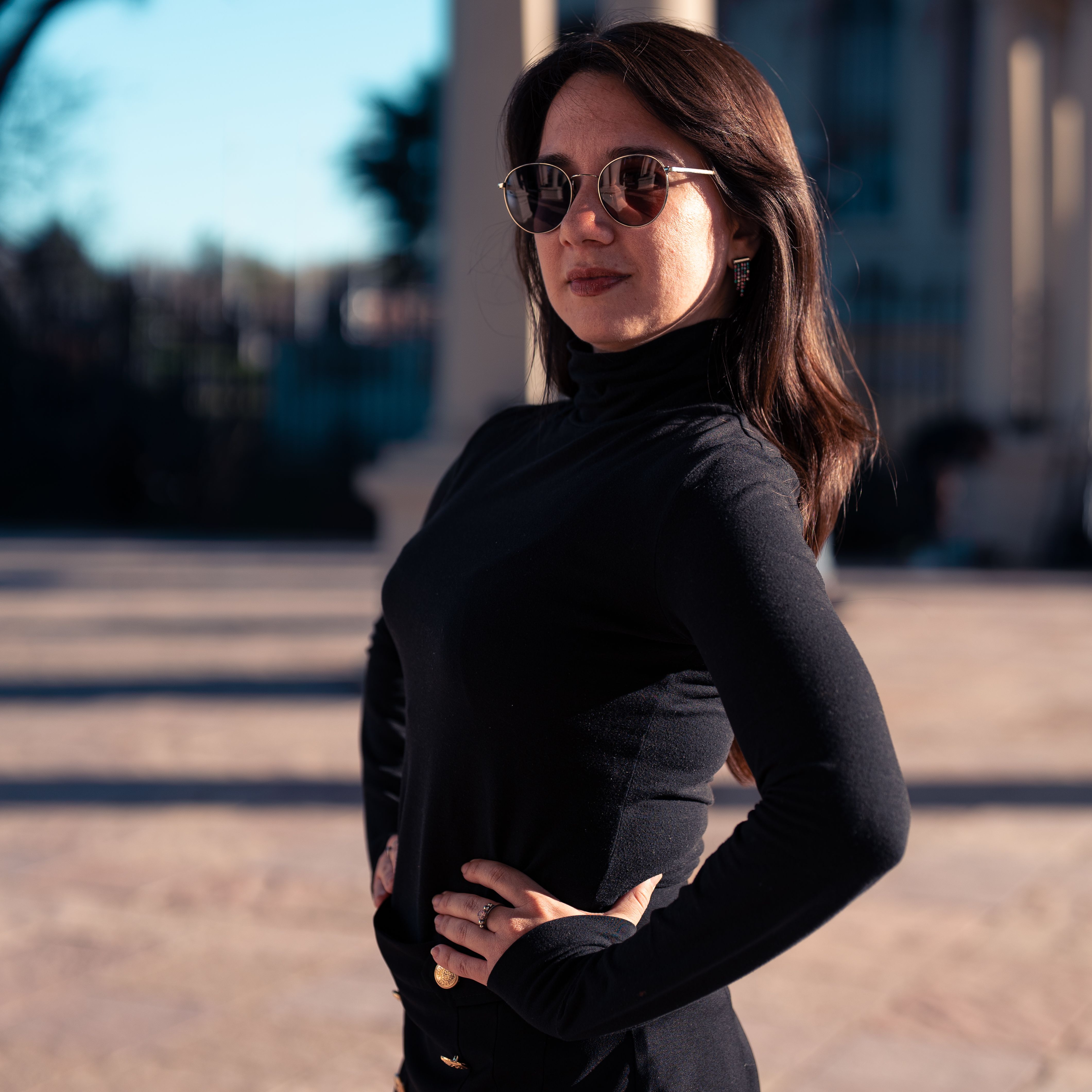 Confident woman in sunglasses and black outfit posing in sunlight outdoors.