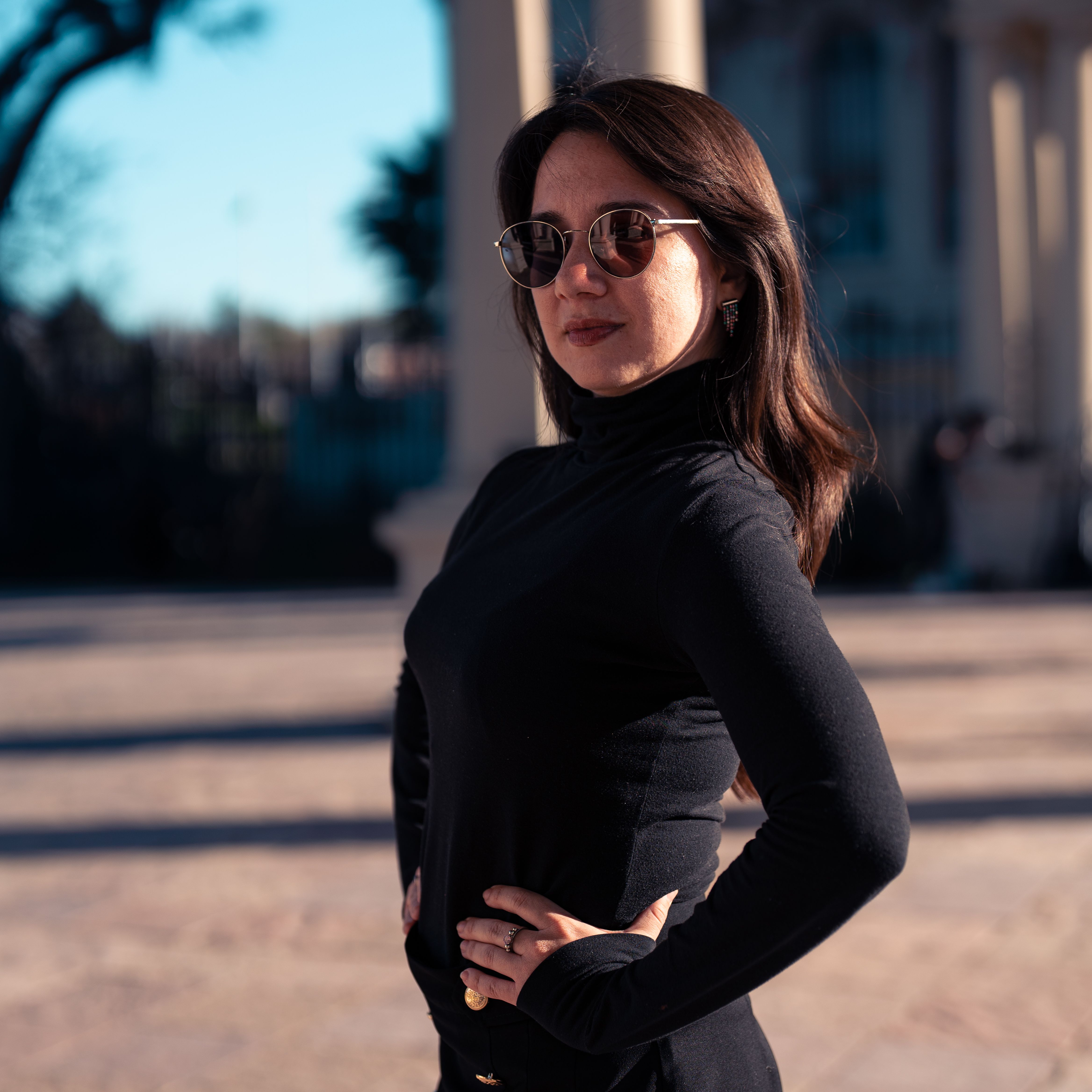 Confident woman in black clothing poses outdoors wearing sunglasses.