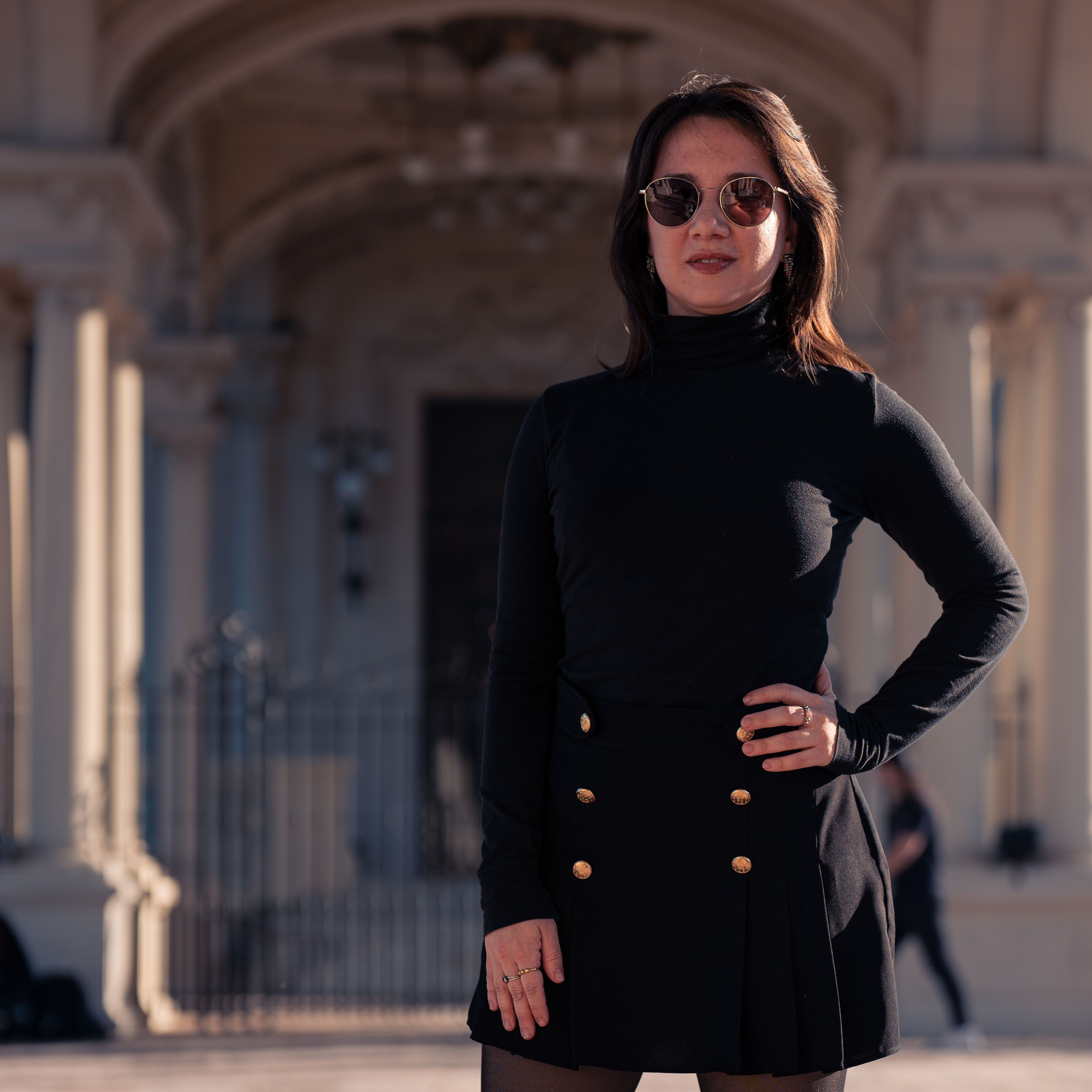Confident woman wearing a black outfit poses outdoors with a historical backdrop.
