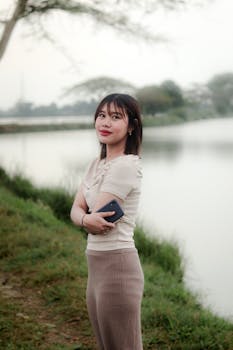 Smiling young adult woman holding a phone, standing by a peaceful lake, embracing nature.