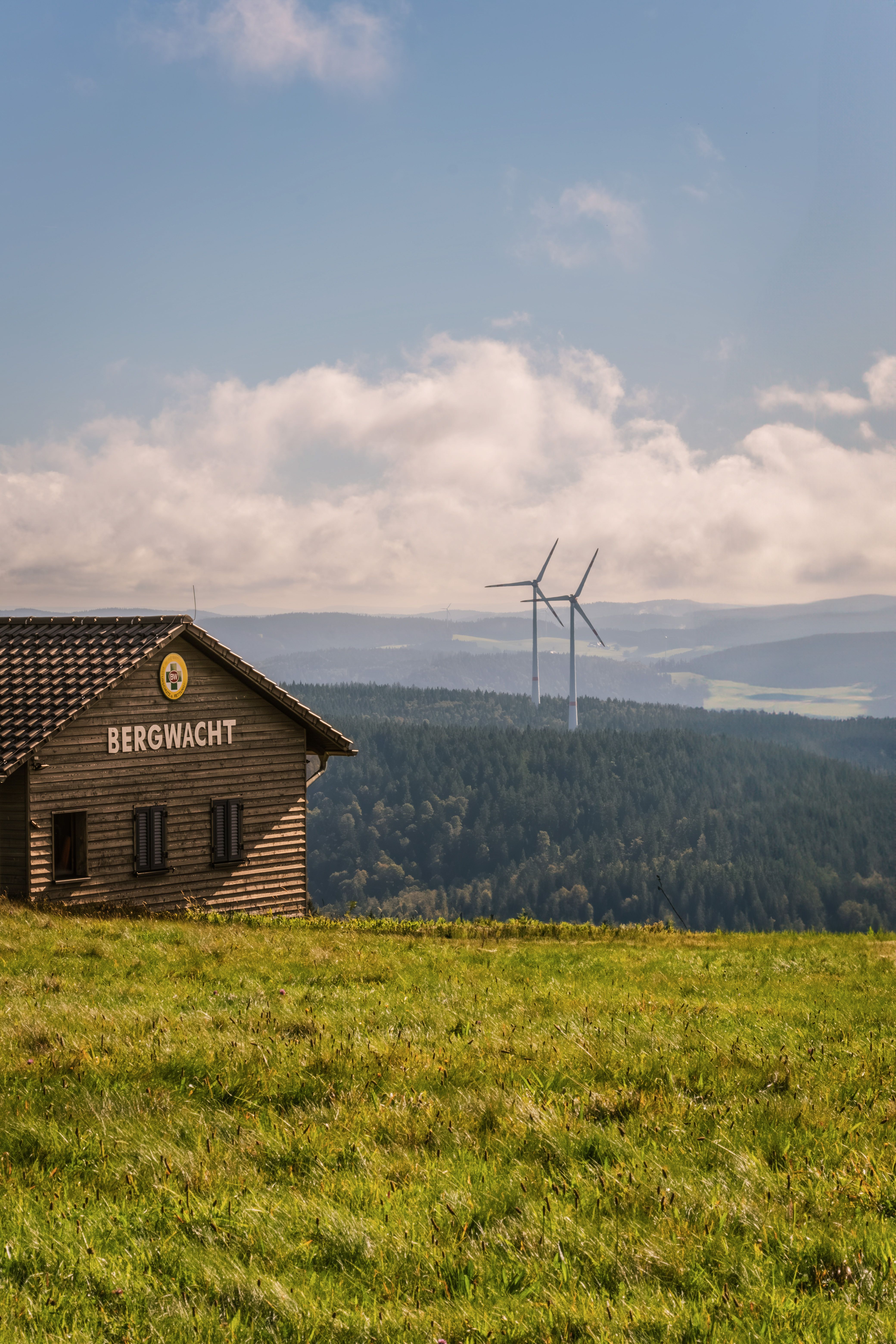 Wooden cabin in a hilly landscape with wind turbines in the background.