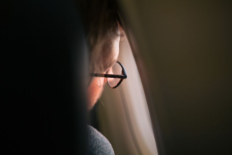 A thoughtful man gazing out an airplane window, wearing eyeglasses, in soft ambient lighting.