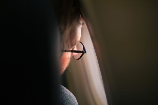 A thoughtful man gazing out an airplane window, wearing eyeglasses, in soft ambient lighting.