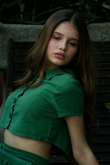 A young woman poses gracefully in a green outfit against a lush outdoor backdrop, showcasing elegance and style.
