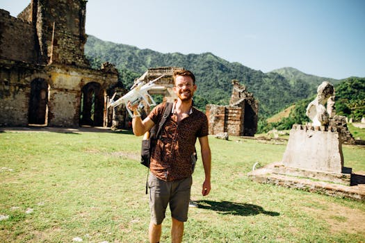 A stylish man poses with a drone at historic ruins in a mountainous landscape.
