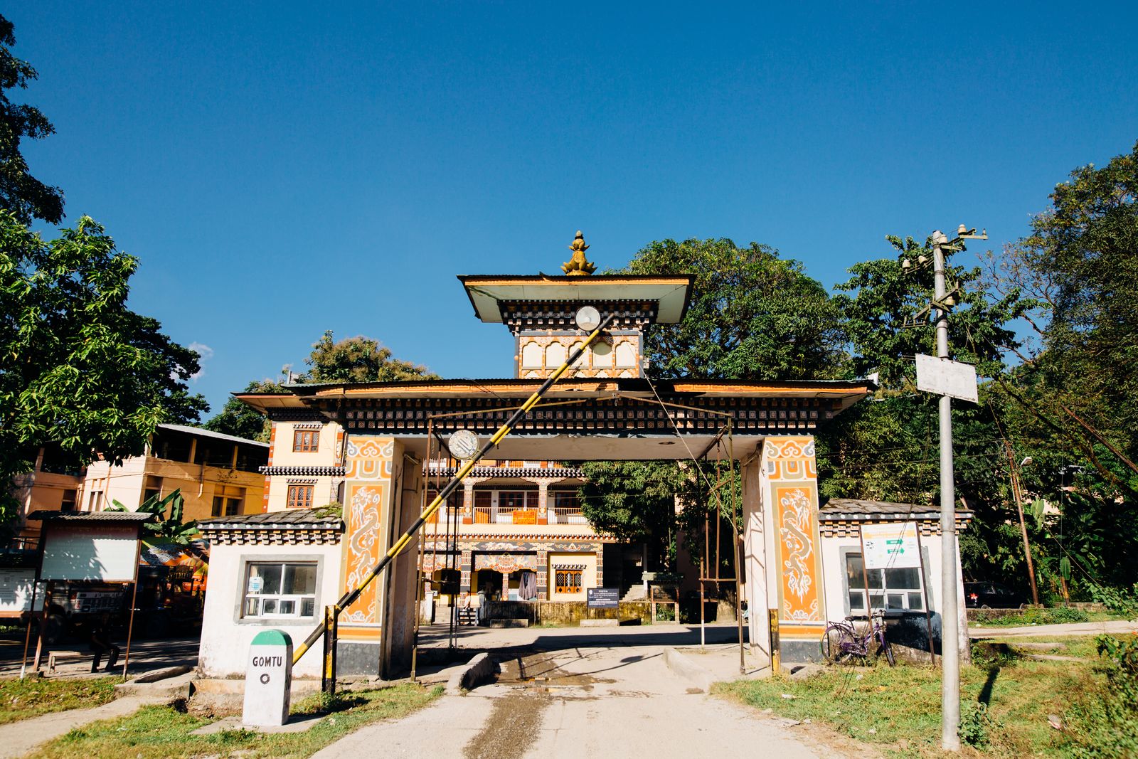 A painted Bhutanese gate arching over the road at a checkpost, sentry booths and green hills under a clear sky