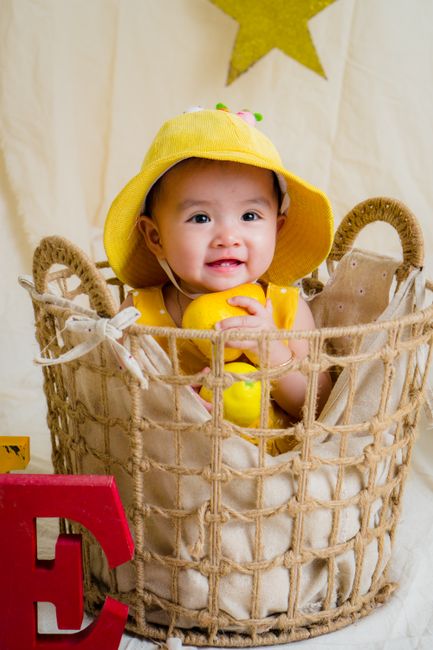 Cute baby in yellow hat smiling joyfully inside a woven basket with toys.