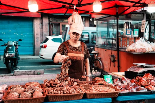 A vendor at a street market selling various meats under a red tent with urban background.