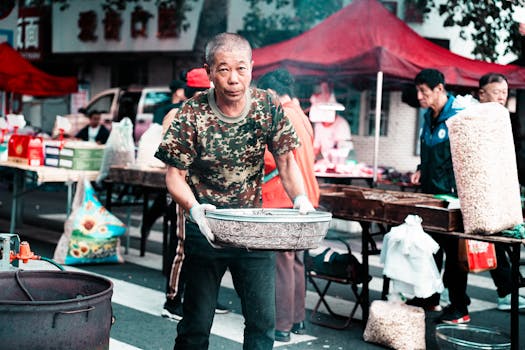 Man selling roasted seeds at an outdoor market with other vendors.