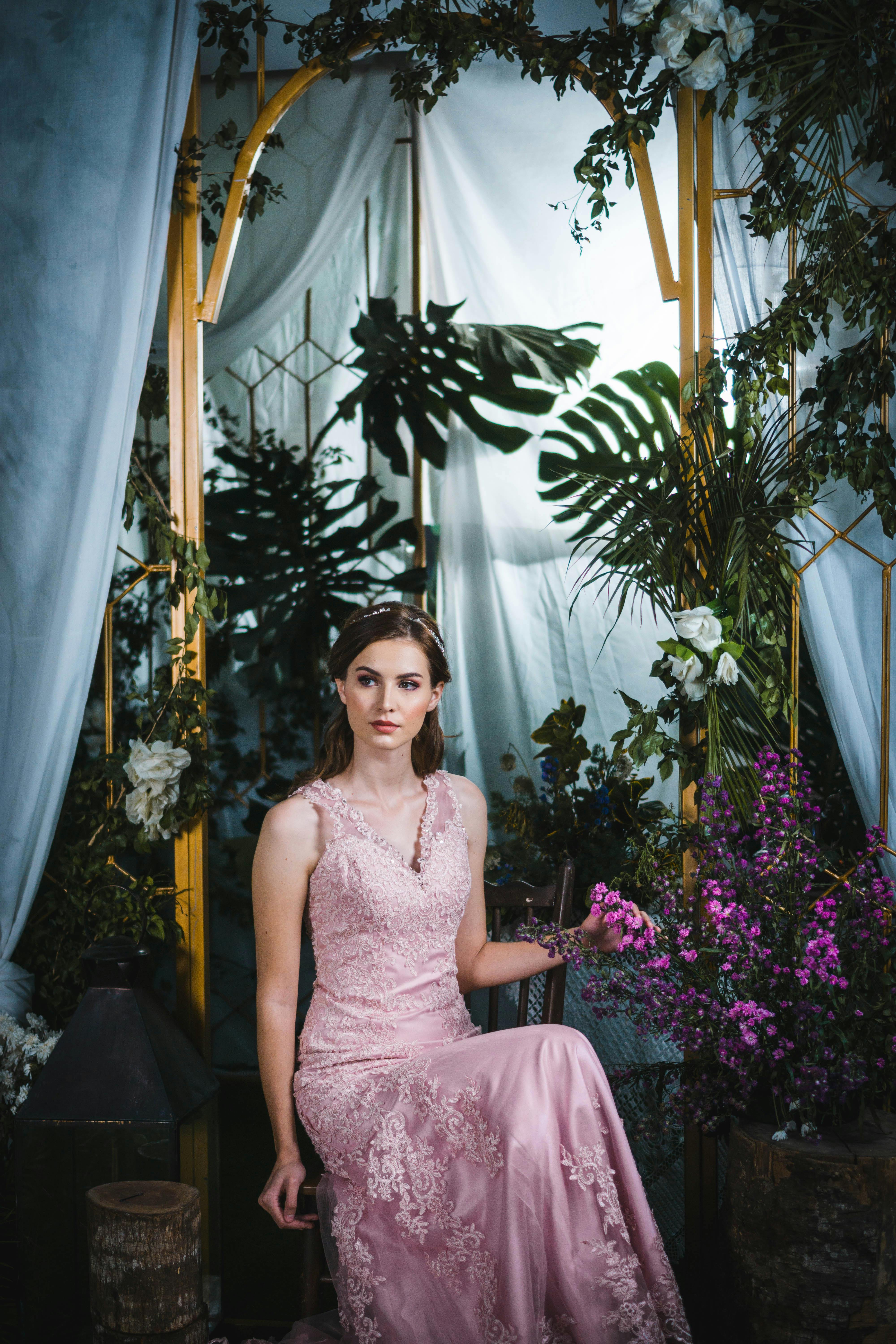 一位年轻女子身穿粉色蕾丝连衣裙，在室内的绿植和花卉装饰间优雅地摆着姿势