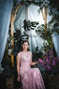 一位年轻女子身穿粉色蕾丝连衣裙，在室内的绿植和花卉装饰间优雅地摆着姿势