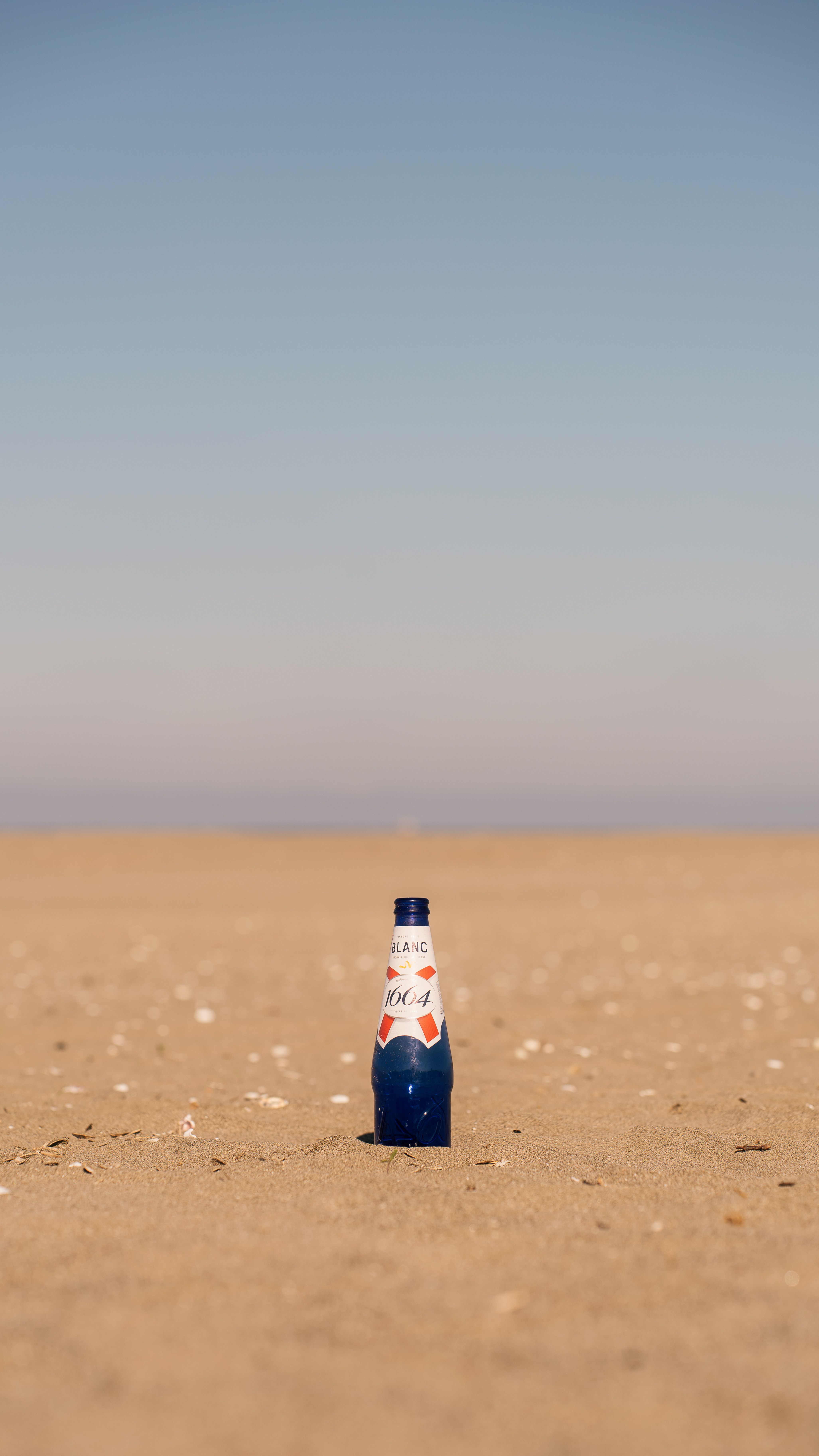 A blue bottle stands on a sandy beach under a clear sky, capturing minimalism and solitude.