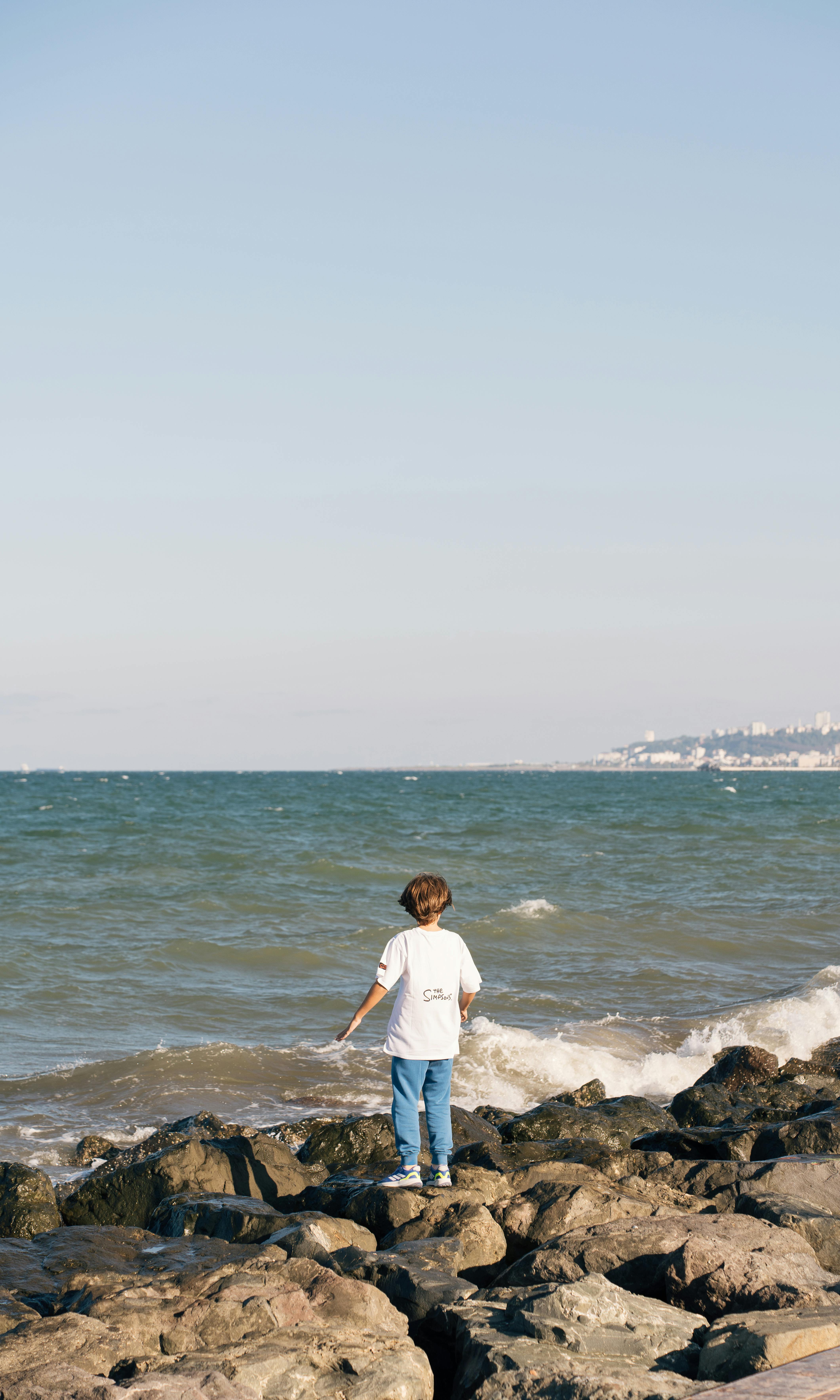 A child stands on coastal rocks, gazing at the ocean with a distant city view.