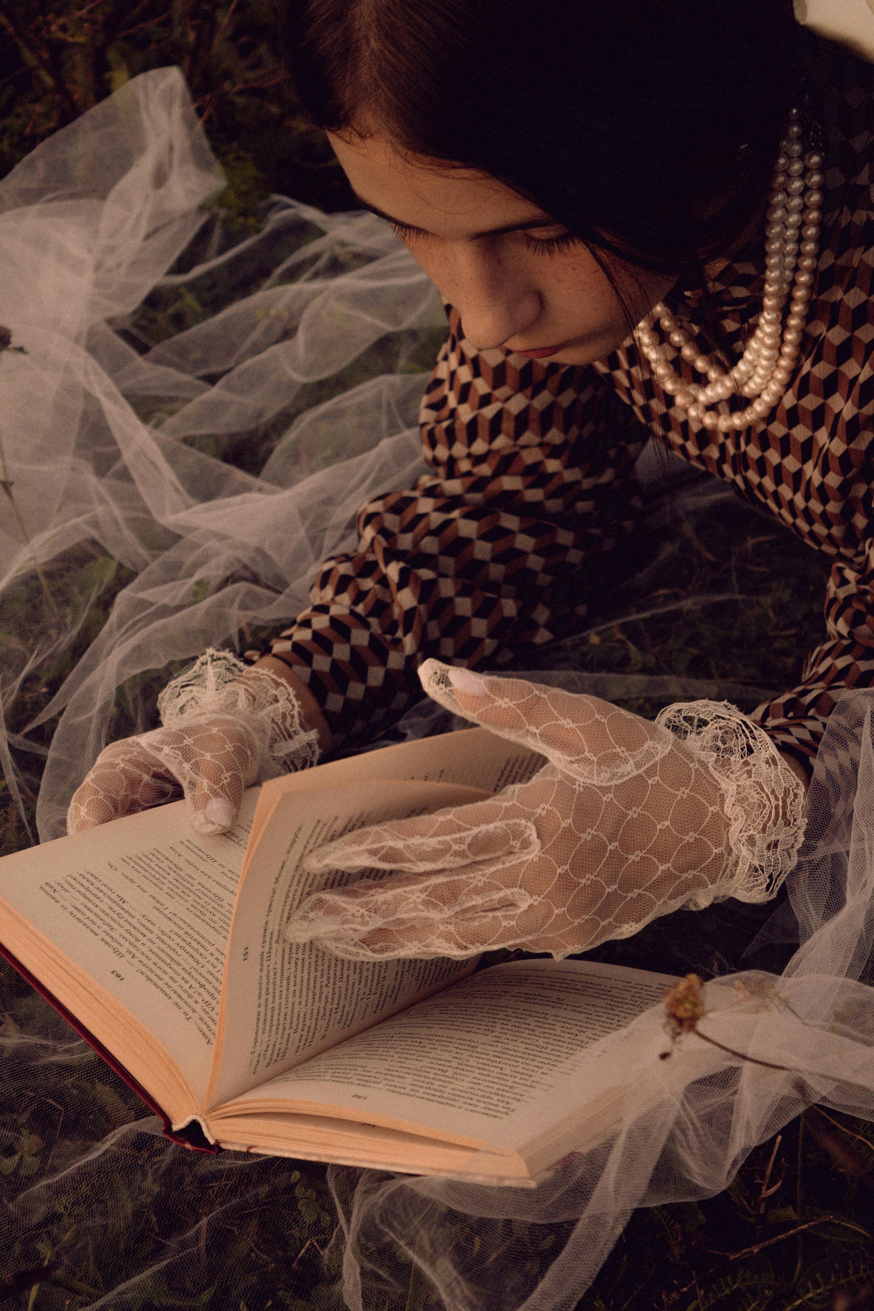 Woman in vintage attire with lace gloves reads a book outdoors.