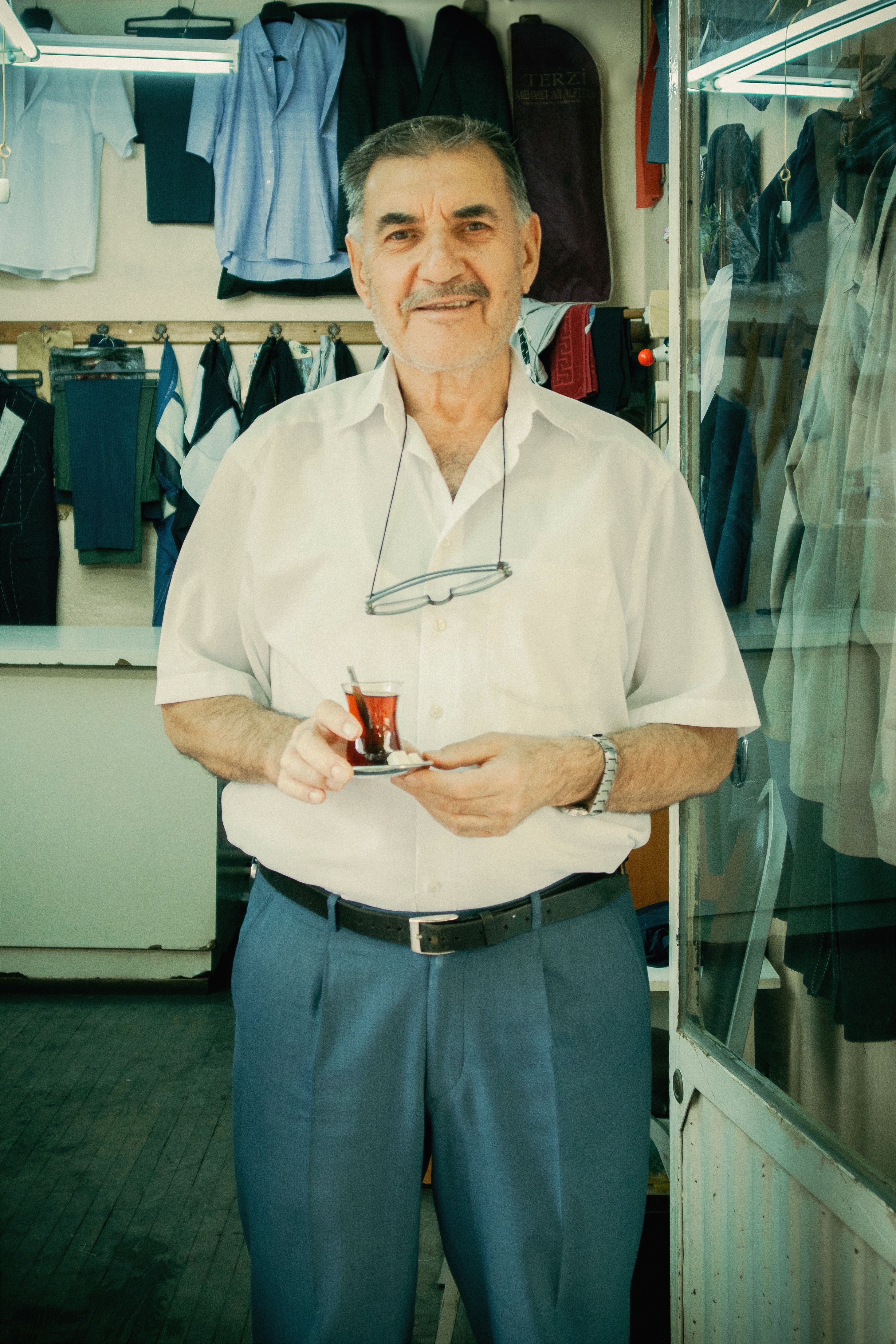 Senior man holding a traditional tea glass in a clothing shop, smiling warmly at the camera.