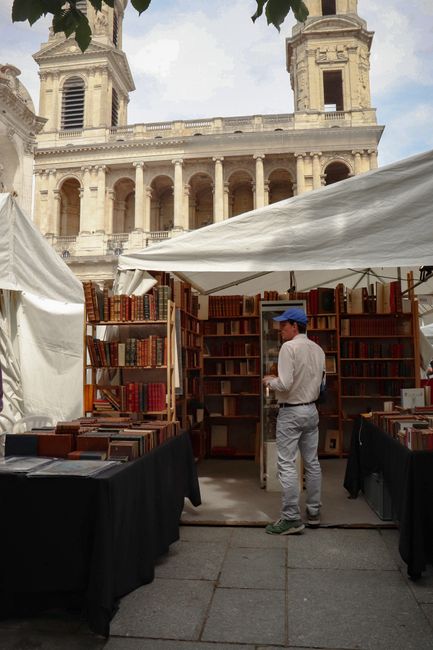 A man browsing vintage books in a Paris outdoor market with Saint-Sulpice Church in the background.