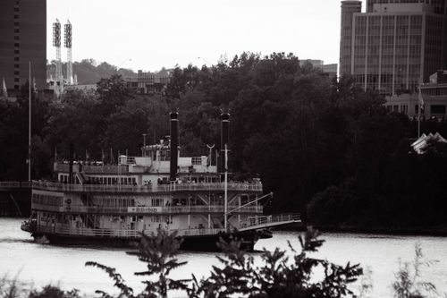 Free A traditional riverboat sails along a river with a city skyline and lush trees in the background. Stock Photo