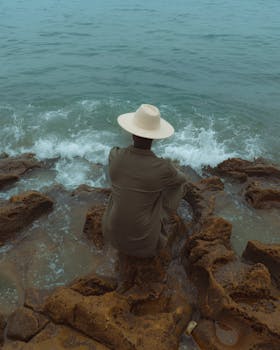 A solitary figure with a hat gazes at the ocean, surrounded by rugged rocks, evoking tranquility and introspection.