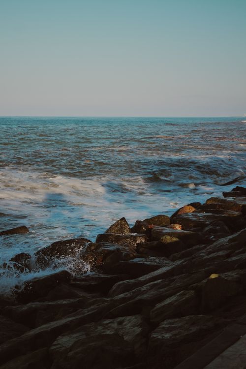 Free A tranquil scene of ocean waves crashing against a rugged rocky coastline under a clear sky. Stock Photo
