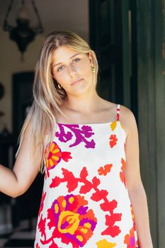 A young woman in a vibrant floral dress stands at the entrance of a house, enjoying a sunny day.