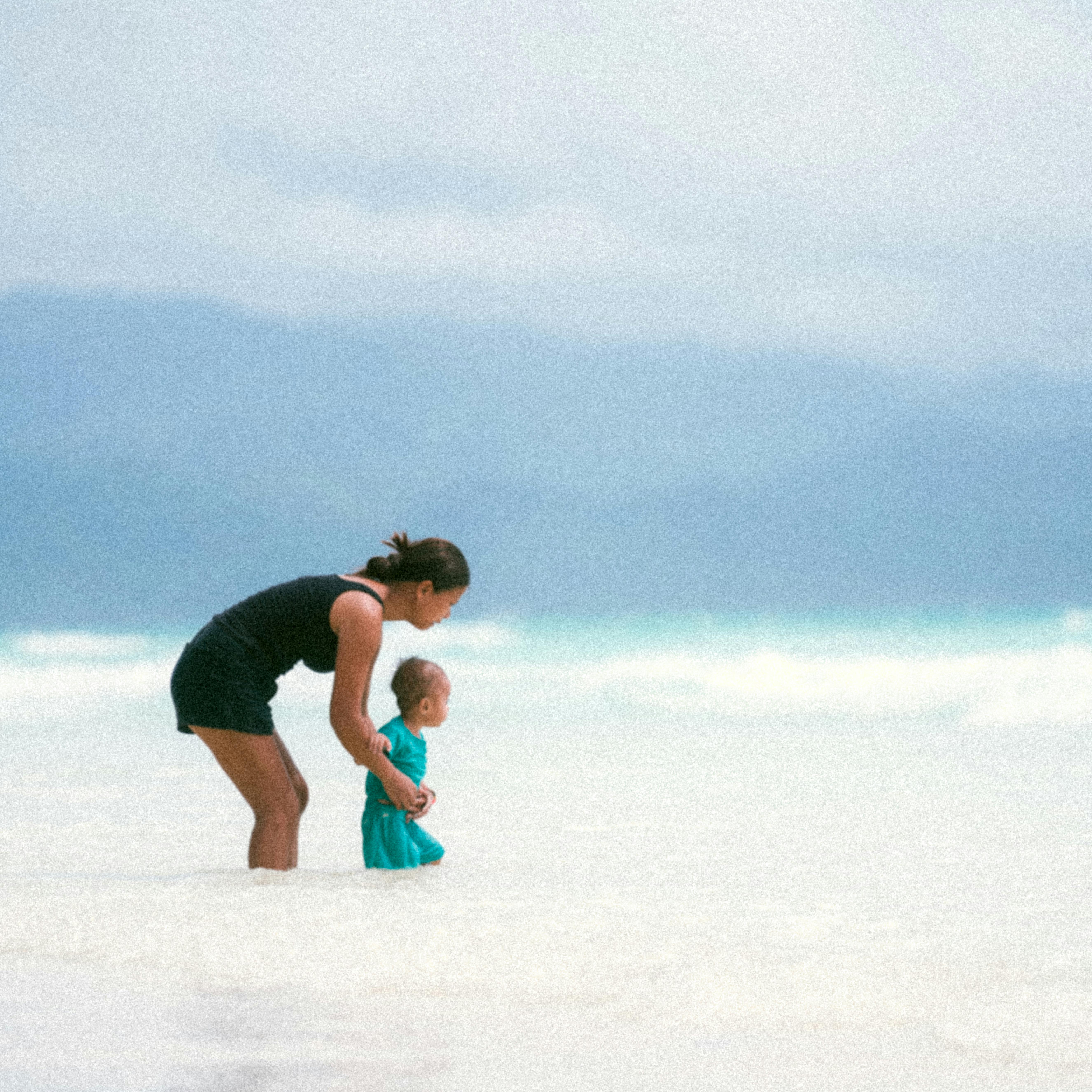 A young child sitting on a sandy beach playing with a blue bucket.