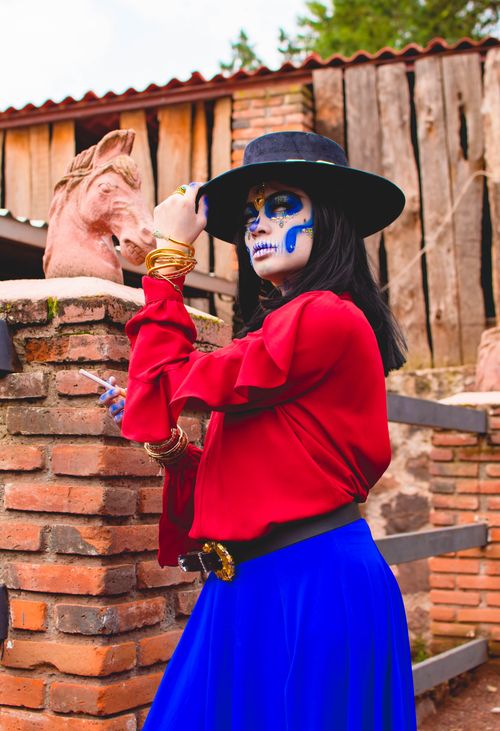 Free Colorful portrait of a woman wearing traditional Catrina makeup, a black hat, and vibrant attire outdoors, celebrating Mexican culture. Stock Photo