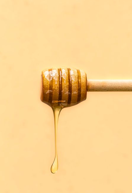 Close-up of honey drizzling from a wooden dipper against a soft yellow background.