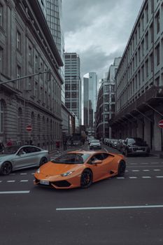 A vibrant orange Lamborghini drives through the bustling streets of Frankfurt, Germany.