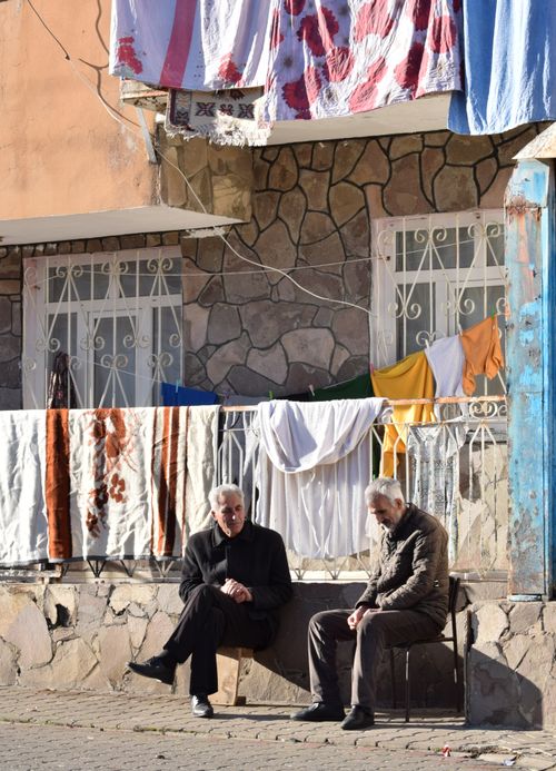 Free Two elderly men sitting outside a stone building with laundry hanging, enjoying a sunny day. Stock Photo