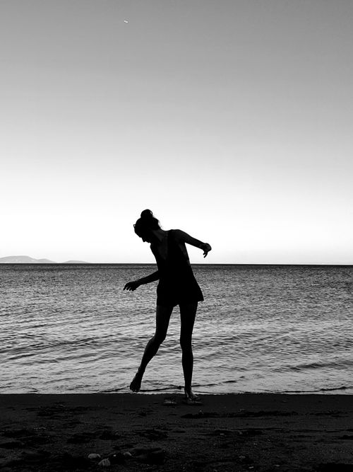 Free A silhouette of a woman standing on a tranquil Greek beach at sunset, captured in monochrome. Stock Photo