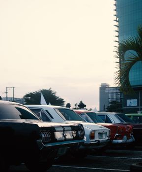 Classic cars lined up in a city parking lot, captured at sunrise, with modern buildings in the backdrop.