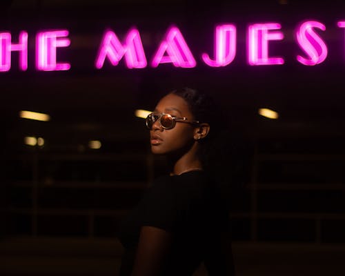 Selective Focus Photo of Woman in Black Top and Sunglasses Posing with Neon Sign in the Background