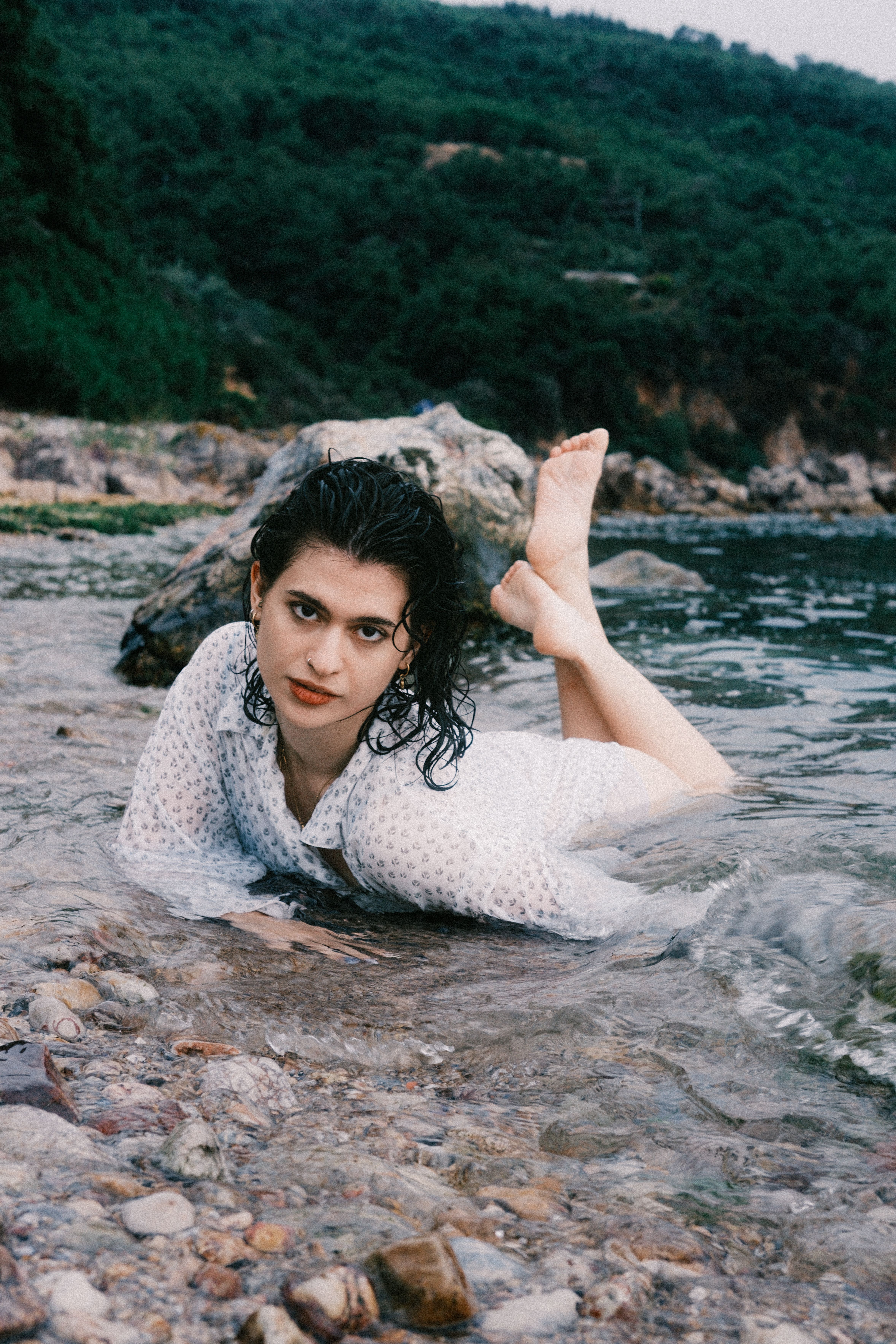 A woman in a white dress poses in the water on a rocky beach, surrounded by lush greenery.