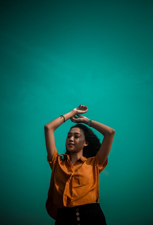 Happy woman in orange top raising her arms above her head against a teal background - fitness lifestyle benefits