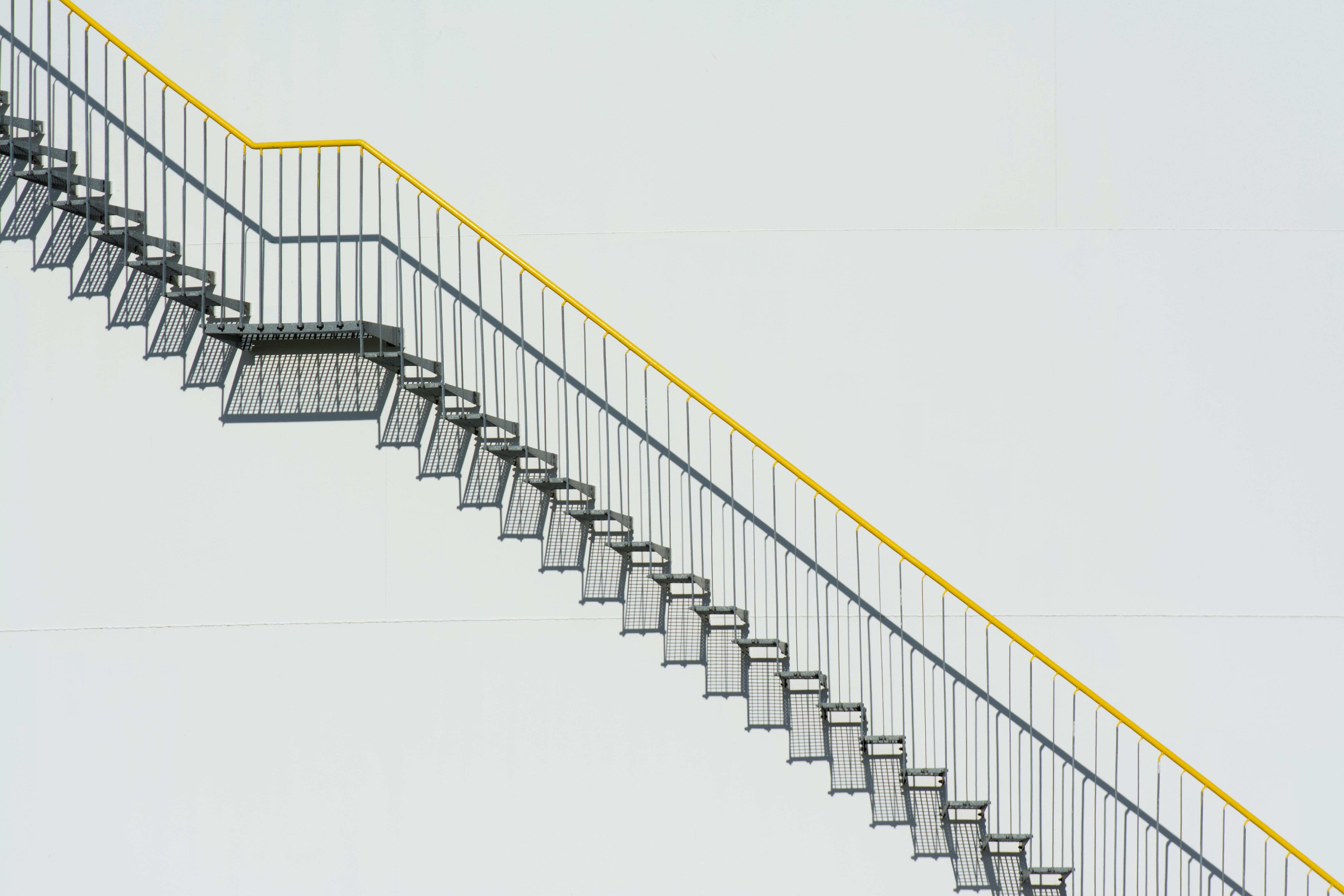 A modern minimalist stairway with yellow railing diagonally ascends a plain white wall.
