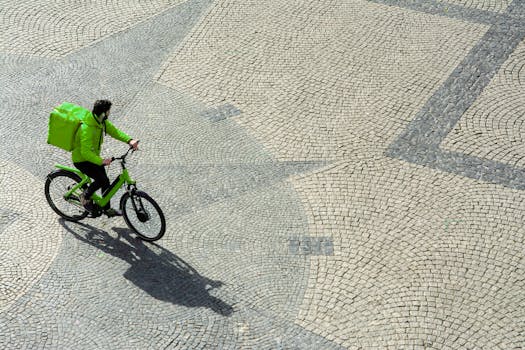 Courier cyclist rides a bicycle with a green backpack across a sunlit cobbled street, casting a long shadow.