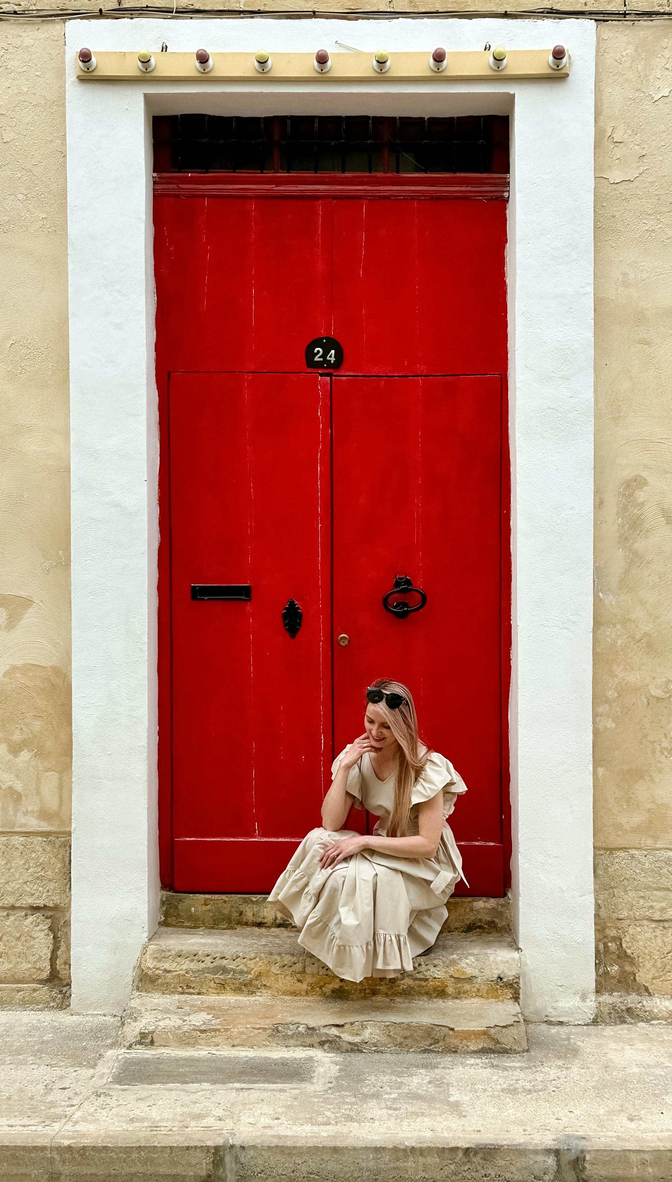 A woman in a beige dress sits on stone steps in front of a vibrant red door in Siġġiewi, Malta.
