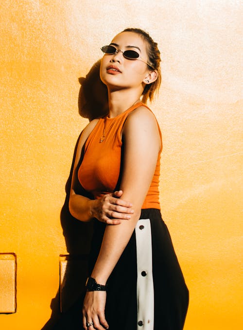 Photo of Woman in Sunglasses,Orange Top, and Black Bottoms Standing Beside Yellow Wall