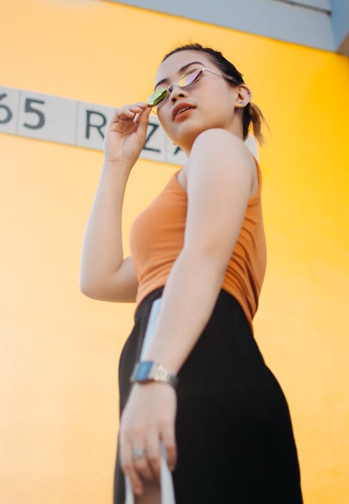 Free Low Angle Photo of Woman in Sunglasses,Orange Top, and Black Bottoms Standing Beside Yellow Wall Stock Photo