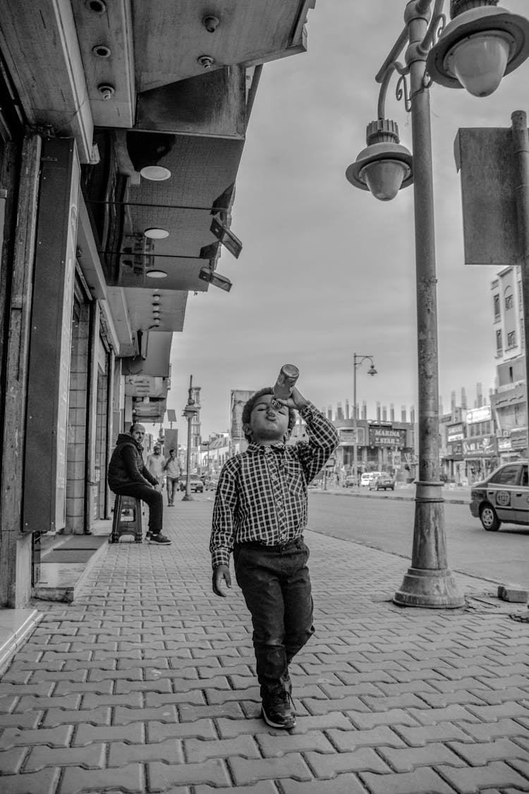 Grayscale Photo Of Boy In Dress Shirt Walking On Side Walk While Drinking From A Glass Bottle